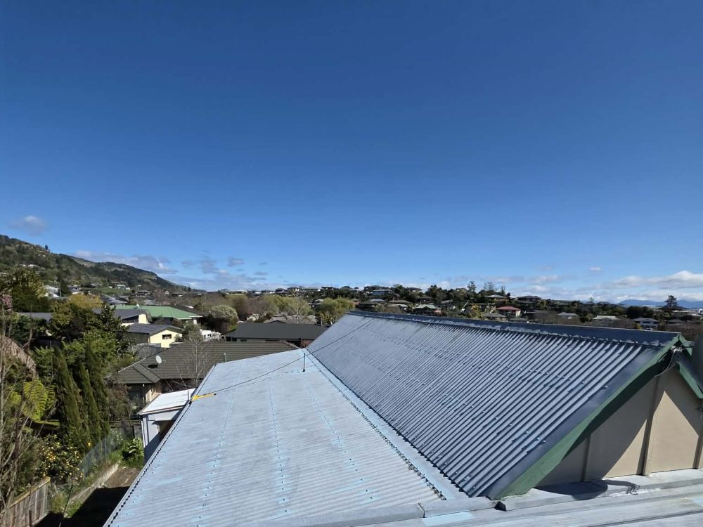 A tired grey and dark corrugated iron roof seen from above, with houses and hills behind
